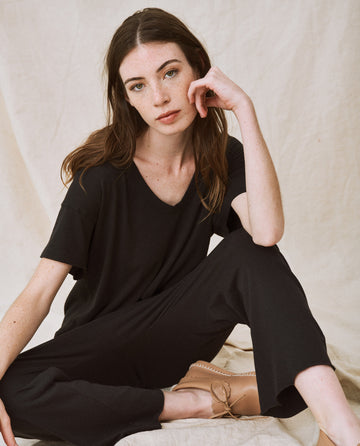 A woman with long brown hair is sitting cross-legged on the floor, resting her chin on her hand. She is wearing The Jersey Crop in Almost Black from THE GREAT. CORE KNITS and beige shoes, maintaining a neutral expression.