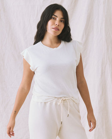 Person wearing a THE GREAT. FALL 22 KNITS The Peak Shoulder Tee in Washed White and high-waisted denim standing in front of a cream-colored backdrop.