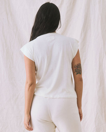A person with long dark hair, seen from behind, is wearing The Peak Shoulder Tee in Washed White from THE GREAT. FALL 22 KNITS and light-colored high-waisted denim pants, with a tattoo visible on their right arm.