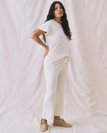 A person wearing "The Slim Tee. Solid -- Washed White" by THE GREAT. CORE KNITS and matching pants stands against a white fabric background, looking at the camera with a slight smile. They have long dark hair and are wearing tan shoes.