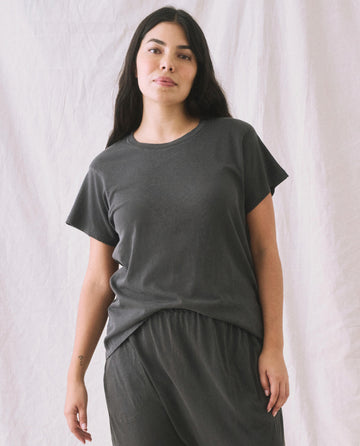A person with long dark hair is wearing THE GREAT. CORE KNITS' The Slim Tee in Washed Black, paired with matching pants, standing against a plain white backdrop.