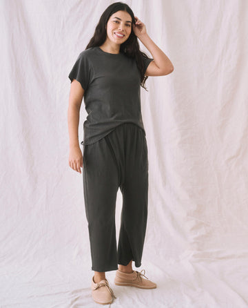 A woman with long dark hair smiles and poses, wearing The Slim Tee in Washed Black by THE GREAT. CORE KNITS and pants, complemented by beige shoes, against a white draped background.
