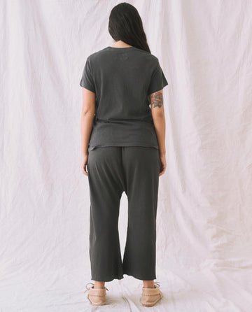 A person stands with their back to the camera against a white backdrop, wearing THE GREAT. CORE KNITS' The Slim Tee in Washed Black and pants, with an arm tattoo visible.