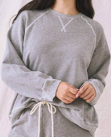 Close-up of a person wearing The Slouch Sweatshirt in Varsity Grey by THE GREAT. CORE KNITS, featuring raglan sleeves, fuller sleeves, and a drawstring waist, standing against a neutral background.