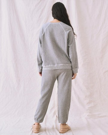 A person with long hair is facing away from the camera, wearing The Slouch Sweatshirt by THE GREAT. CORE KNITS in Varsity Grey, along with matching grey sweatpants and tan moccasin-style slippers, standing against a light-colored fabric backdrop.