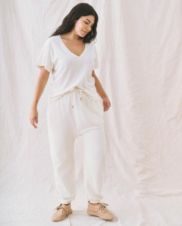 Person standing wearing The V-Neck Tee in Washed White from THE GREAT. SP22 APRILCAP paired with beige shoes, against a light fabric backdrop, featuring a boxy fit that adds a touch of casual elegance.