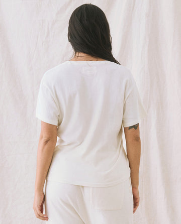 A person with long dark hair is standing with their back to the camera, wearing The V-Neck Tee in Washed White from THE GREAT. SP22 APRILCAP, along with white pants, against a light-colored fabric backdrop.