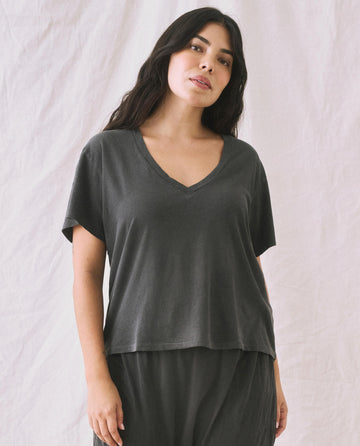 A person with long dark hair stands against a plain backdrop wearing a boxy fit, Washed Black V-Neck Tee and matching pants by THE GREAT. SP22 APRILCAP.