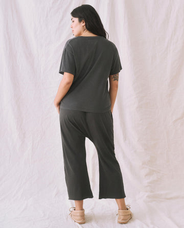 Person wearing The V-Neck Tee in Washed Black by THE GREAT. SP22 APRILCAP and loose-fitting pants stands barefoot on a crumpled white sheet, facing away from the camera.
