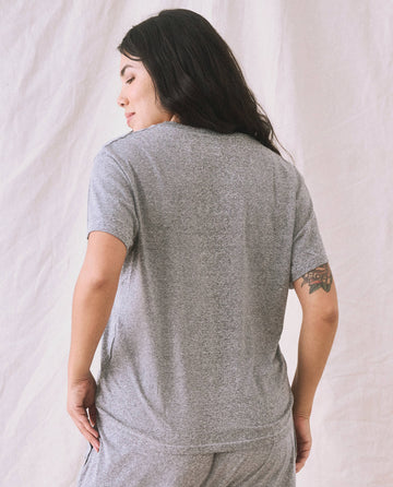 A person with long dark hair and a tattoo on their left arm is standing with their back turned, wearing The V-Neck Tee. in Heather Grey by THE GREAT. SP22 APRILCAP, with a boxy fit and matching pants against a light fabric backdrop.