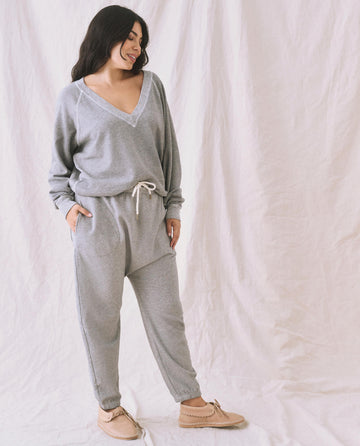 A person stands against a draped fabric background, wearing the Varsity Grey V-Neck Sweatshirt from THE GREAT. FALL 24 KNITS and matching pants with hands in pockets, paired with beige shoes.
