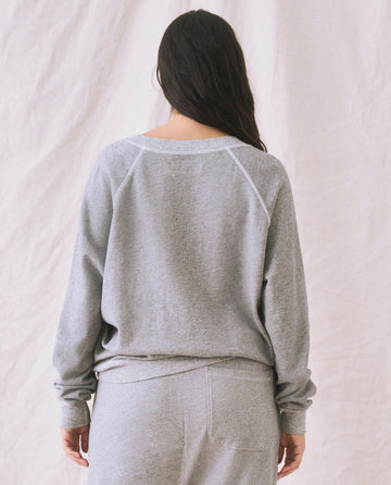 A person with long dark hair is standing facing away, wearing "The V-Neck Sweatshirt" in Varsity Grey from THE GREAT. FALL 24 KNITS collection, along with matching pants, against a neutral background.