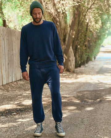 A man wearing a blue sweatshirt, **The Men's Stadium Sweatpant in True Navy by THE GREAT. HOL 22 MEN**, and a green beanie walks on a dirt path lined with trees and a wooden fence.