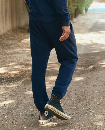 A person clad in The Men's Stadium Sweatpant in True Navy from THE GREAT. HOL 22 MEN, paired with black high-top sneakers, is walking on a dirt path. The individual is captured from the back.