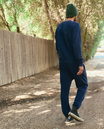 A person wearing The Men's Stadium Sweatpant in True Navy from THE GREAT. HOL 22 MEN and a green beanie walks down a tree-lined dirt path next to a wooden fence.