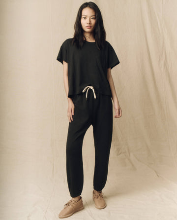 A person stands against a plain backdrop, wearing The Pocket Tee in Solid -- Almost Black from THE GREAT. SP23 CORE KNITS, paired with pants featuring beige drawstrings and beige shoes.