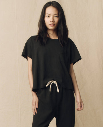 A woman with long dark hair wearing The Pocket Tee in Almost Black by THE GREAT. SP23 CORE KNITS and black drawstring pants, standing in front of a beige background.