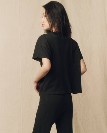 A woman with loose, dark hair is standing, turned slightly to the side, wearing The Pocket Tee by THE GREAT. SP23 CORE KNITS in Solid -- Almost Black and black pants against a beige background.