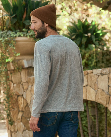 A man wearing The Men's Boxy Henley in Heather Grey by THE GREAT. HOL 22 MEN, a brown beanie, and blue jeans is walking outdoors near a stone wall and greenery in Los Angeles. His back is turned to the camera.