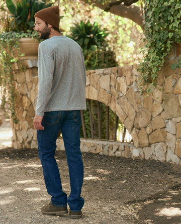 A man in The Men's Boxy Henley - Heather Grey from THE GREAT. HOL 22 MEN, along with a beanie, jeans, and brown shoes, stands outdoors near a stone wall with plants in Los Angeles.