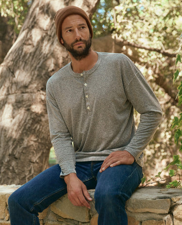 A bearded man wearing a brown beanie, The Men's Boxy Henley in Heather Grey by THE GREAT. HOL 22 MEN, and blue jeans sits on a stone ledge in a forested area.