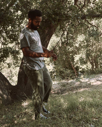 A person wearing The Men's Boxy Crew in Heather Grey by THE GREAT. MAN, green pants, and black sneakers stands in a wooded area with sunlight filtering through the trees.
