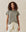 A person with curly hair wearing THE GREAT. CORE KNITS' The Slim Tee in Heather Grey and white drawstring pants stands against a beige fabric background.