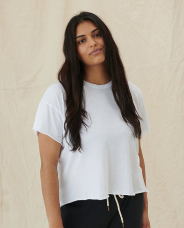 A person with long, dark hair wears THE GREAT. CORE KNITS' super soft cotton cropped top, The Crop Tee in TRUE WHITE, and stands in front of a beige background.