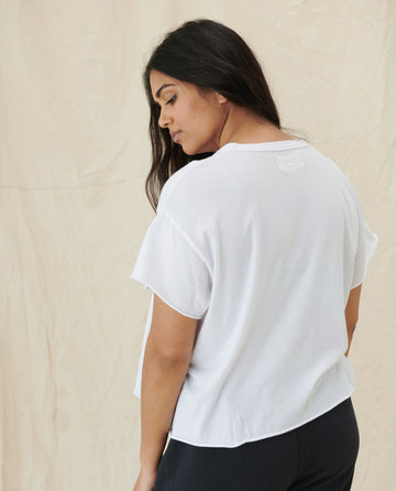 A person with long dark hair wearing 'The Crop Tee' in TRUE WHITE from THE GREAT. CORE KNITS and black pants is standing with their back turned, looking over their shoulder. The background is a plain beige curtain.