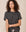 A woman with curly hair, wearing The Crop Tee in Washed Black from THE GREAT. CORE KNITS and high-rise jeans made from super soft cotton, stands against a plain background, looking to her left.