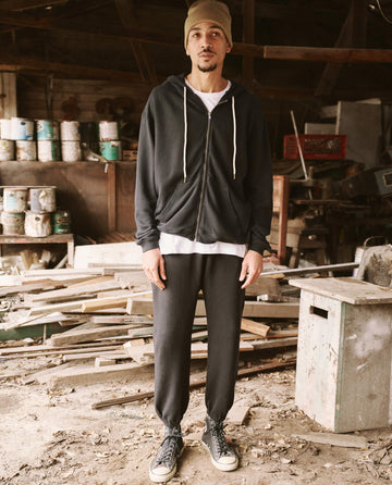 A person stands in a cluttered workshop wearing THE GREAT. SP22 D2 MENS The Men's Zip Up Hoodie in Almost Black, matching sweatpants, a beige beanie, and black sneakers. The ground is dusty and scattered with wooden planks.
