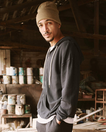 A man wearing a beige beanie and "The Men's Zip Up Hoodie -- Almost Black" by THE GREAT. SP22 D2 MENS stands in a cluttered workshop with cans and tools in the background, showcasing his versatile layers.