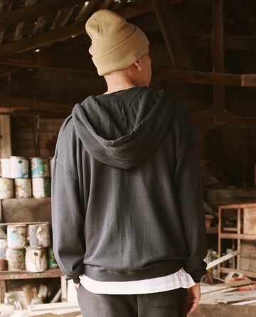 A person wearing a beige beanie and The Men's Zip Up Hoodie in Almost Black by THE GREAT. SP22 D2 MENS is standing indoors with their back to the camera, in a setting that includes paint cans and wooden structures.