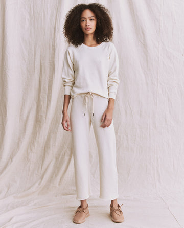 A person with curly hair stands against a cream-colored background, wearing The Shrunken Sweatshirt in Washed White from THE GREAT. CORE KNITS, paired with matching drawstring pants made of 100% cotton and light brown shoes.