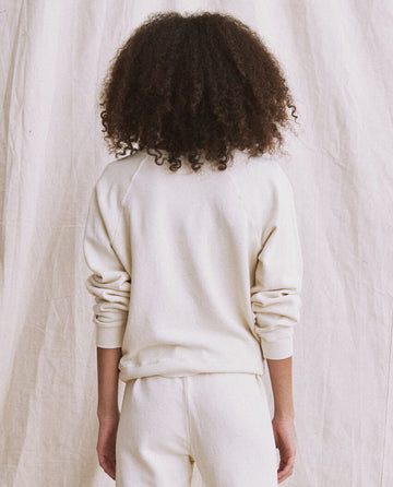 A person with curly hair stands with their back to the camera, wearing The Shrunken Sweatshirt in Washed White by THE GREAT. CORE KNITS and matching pants against a light fabric background.