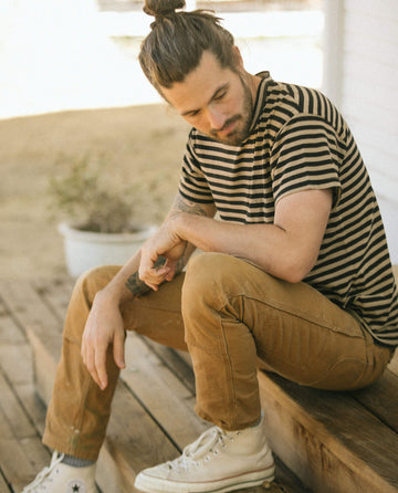 A man with a bun hairstyle and beard, wearing "The Men's Boxy Crew -- Black and Camel Stripe" by THE GREAT. SU22 MENS, sits on wooden steps outdoors, looking down pensively.