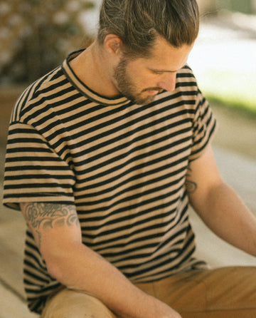 A man with long hair in a bun, wearing The Men's Boxy Crew in Black and Camel Stripe from THE GREAT. SU22 MENS, looks down. He has tattoos on his left arm and is sitting outdoors.