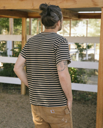 A person with a man bun and "The Men's Boxy Crew" in Black and Camel Stripe by THE GREAT. SU22 MENS stands outdoors with hands in pockets, facing away from the camera. Tattoos are visible on their arm.