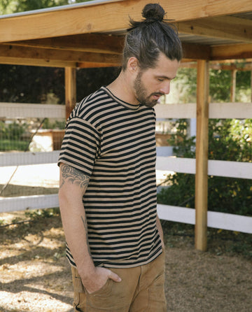 A man with a beard and a top knot hairstyle, sporting The Men's Boxy Crew in Black and Camel Stripe by THE GREAT. SU22 MENS, paired with brown pants, stands outdoors near a wooden structure and white fence.