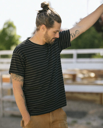 A man with his hair in a bun, wearing The Men's Boxy Crew in Black and Charcoal Stripe from THE GREAT. SU22 MENS, and brown pants, stands near a white fence with his right arm raised. This comfortable look is perfect for any casual outing, and the tee is proudly made in the U.S.A.