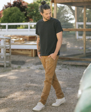 A man with long hair in a bun, wearing The Men's Boxy Crew in Black and Charcoal Stripe by THE GREAT. SU22 MENS, tan pants, and white sneakers, walks outdoors on a gravel path.