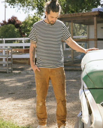 A man with tattoos is standing outdoors by a vintage car, wearing The Men's Boxy Crew in black and cream stripe from THE GREAT. SU22 MENS, paired with brown pants, with his hands in his pockets.