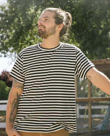 A man with a beard and tattoos, wearing The Men's Boxy Crew in Black and Cream Stripe from THE GREAT. SU22 MENS—a classic staple in any wardrobe—standing outdoors with a tree in the background.
