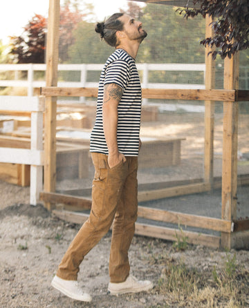 A man in a classic staple, The Men's Boxy Crew in Black and Cream Stripe by THE GREAT. SU22 MENS, paired with brown pants and white sneakers, stands outside looking up with his hands in his pockets. His casual wardrobe choice is complemented by the wooden fence and greenery in the background.