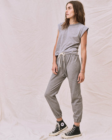 A woman stands against a plain fabric backdrop, wearing The Peak Shoulder Tee in heather grey by THE GREAT. FALL 22 KNITS and matching drawstring joggers, complemented by black high-top sneakers. She looks to her left with a relaxed posture.