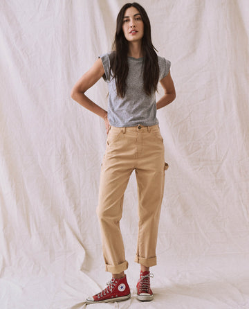 A person stands against a white fabric backdrop, wearing The Peak Shoulder Tee in Heather Grey by THE GREAT. FALL 22 KNITS, beige rolled-up pants, and red high-top sneakers. Their hands are on their hips.
