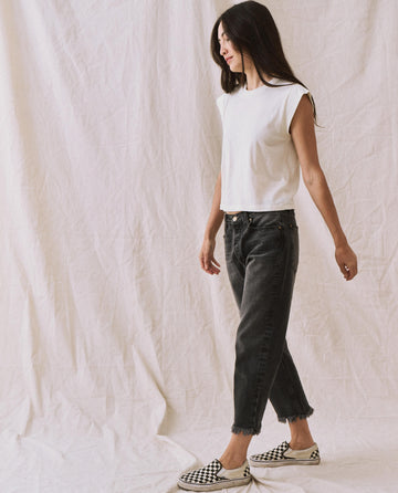 A person with long hair, wearing a handcrafted locally-made Peak Shoulder Tee in washed white from THE GREAT. FALL 22 KNITS, high-waisted denim, and checkered slip-on shoes walks against a neutral fabric backdrop.