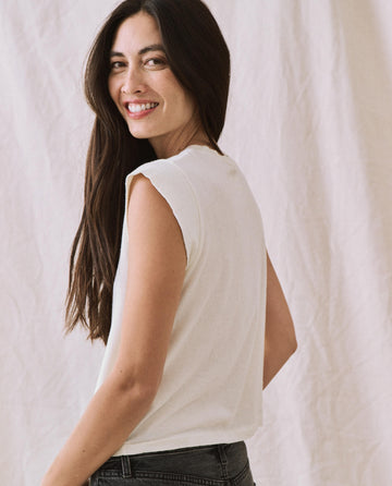 A person with long hair wearing a sleeveless The Peak Shoulder Tee in Washed White from THE GREAT. FALL 22 KNITS smiles while looking over their shoulder, standing against a light-colored fabric backdrop.