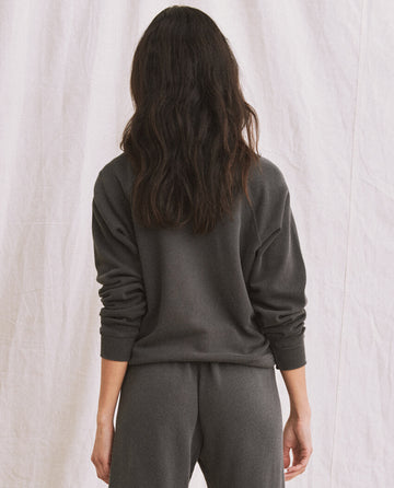 A person with long dark hair wearing The Shrunken Sweatshirt in washed black, standing with their back to the camera against a light-colored background.