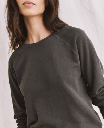 A person with shoulder-length brown hair is wearing The Shrunken Sweatshirt in washed black by THE GREAT. CORE KNITS, standing against a light-colored background. The photo shows their upper body, without revealing their full face.